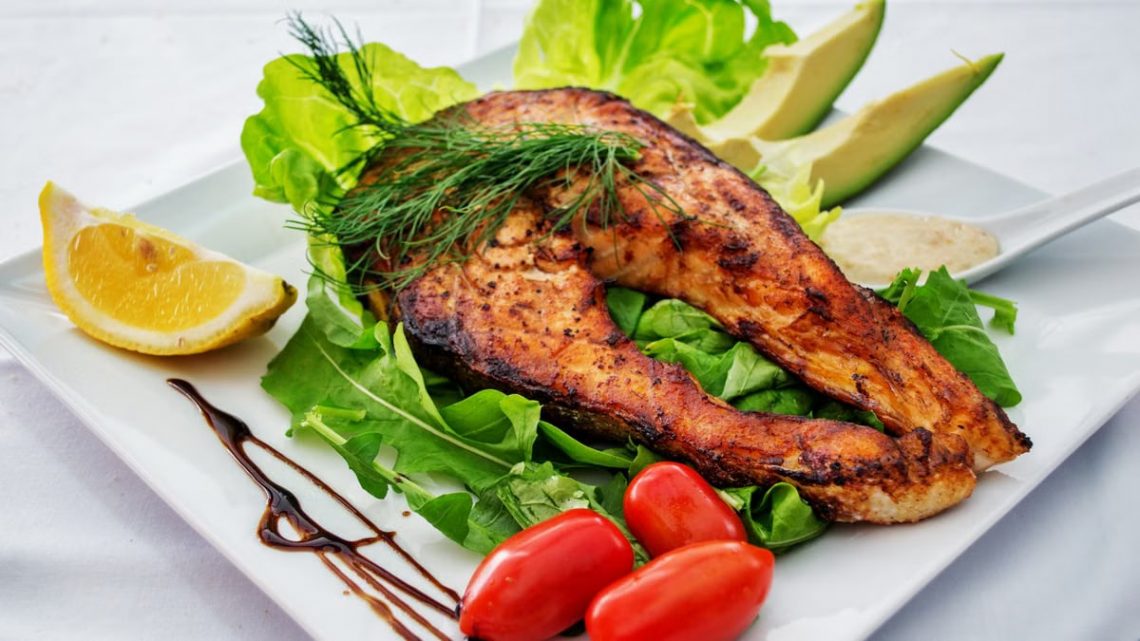Delicious grilled salmon steak served with avocado, lettuce, cherry tomatoes, and lemon slice on a white plate.