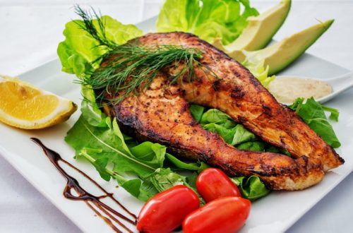 Delicious grilled salmon steak served with avocado, lettuce, cherry tomatoes, and lemon slice on a white plate.